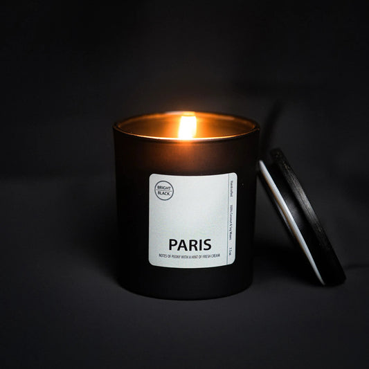 Bright Black Candles in a sleek black frosted glass vessel, being lit to release a luxurious floral blend of peony and gardenia with a hint of French crème brûlée. This 4.75oz candle features a natural wooden wick and offers 70-80 hours of burn time, combining uplifting scents with a modern, minimalistic design.