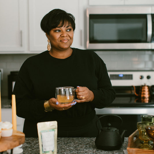 Trista Sanford, founder of Ora Tea House, holding a warm cup of her signature tea in her home. Surrounded by a cozy and inviting atmosphere, she embraces the art of tea-making with passion and care, showcasing the heart behind her handcrafted blends.